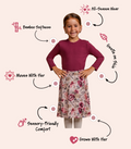 A young girl stands against a light beige background wearing a burgundy long-sleeve top and a floral skirt. Arrows and text call out product features such as bamboo softness, all-season wear, gentle on skin, moves with her, sensory-friendly comfort, and growth with her.