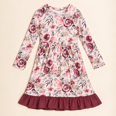 A long-sleeve children's dress with a pink and red floral print on a cream background, featuring a gathered waist and a contrasting dark red ruffled hem, laid flat on a light beige background.