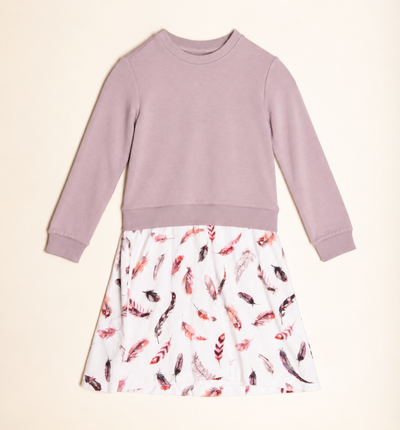 A children's long-sleeve dress laid flat on a warm cream background. The top half is a solid dusty mauve/lavender sweatshirt-style panel, and the bottom half is a white skirt with an allover feather print in pink, red, and dark brown tones.